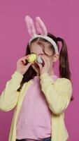 Vertical Video Adorable little girl playing peek a boo game in studio, showing her handcrafted colored easter eggs against pink background. Cheerful playful kid with bunny ears fooling around. Camera A.