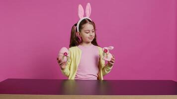 piccolo allegro ragazza mostrando colorato decorato ornamenti su telecamera, la creazione di fatto a mano festivo disposizioni. giovane bambino sensazione eccitato di Pasqua domenica, detiene coniglio e uovo. telecamera b. video