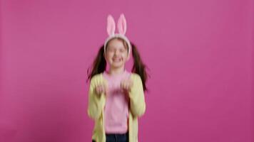 alegre despreocupado Chica de escuela saltando alrededor en estudio, imitando un Conejo y saltando en contra rosado antecedentes. alegre activo niño vistiendo conejito orejas y rebote, adorable niño. cámara b. video