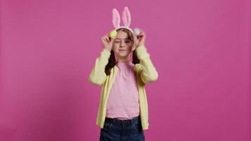aanbiddelijk weinig meisje spelen kijkje een boe spel in studio, tonen haar handgemaakt gekleurde Pasen eieren tegen roze achtergrond. vrolijk speels kind met konijn oren voor de gek houden in de omgeving van. camera b. video