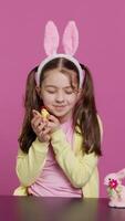 Vertical Video Joyful little girl playing with festive easter decorations in studio, creating arrangements with a chick, rabbit and egg. Smiling cute toddler with bunny ears showing colorful ornaments. Camera A.