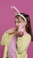 Vertical Video Jolly schoolgirl holding a cute rabbit toy and blowing kisses in studio, being adorable with festive decorations for easter holiday event. Happy child wearing bunny ears and pigtails. Camera A.