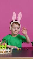 Jeune petit enfant placement lapin oreilles sur le sien tête dans studio, en train de préparer pour Pâques dimanche vacances fête. de bonne humeur peu garçon séance à une table à peindre des œufs et artisanat de fête ornements. caméra b. video