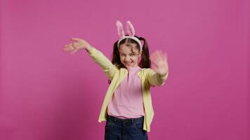 adorabile carino bambino mettendo coniglietto orecchie e agitando a telecamera, godendo Pasqua Domenica celebrazione contro rosa sfondo. sorridente allegro scolara con trecce detto ciao. telecamera b. video