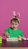 gelukkig kleuter met konijn oren regelen van mand gevulde met geschilderd eieren, creëren feestelijk regelingen voor de Pasen zondag viering. vrolijk weinig kind geniet kunst en ambacht. camera b. video