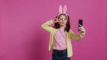 självsäker Lycklig ung flicka tar foton på smartphone webbkamera, känsla söt med henne pigtails och kanin öron. glad leende litet barn tar bilder och lurar runt om i studio. kamera b. video
