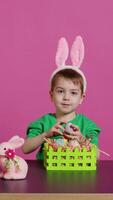 Excited little kid decorating baskets with eggs and grass to prepare for easter holiday festivity, creating festive arrangements. Cute toddler with bunny ears does craft activity. Camera B. video