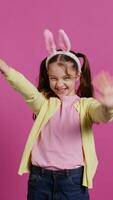 adorable linda niño poniendo conejito orejas y ondulación a cámara, disfrutando Pascua de Resurrección domingo celebracion en contra rosado antecedentes. sonriente alegre Chica de escuela con coletas diciendo Hola. cámara b. video