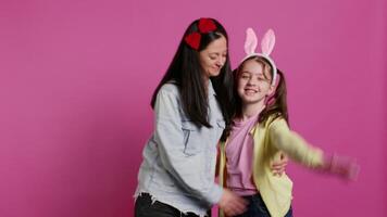 lief schoolmeisje met konijn oren een haar mam golvend Aan camera, hebben pret en lachend tegen roze achtergrond. vrolijk moeder en haar dochter omarmen en zoenen elk ander. camera b. video