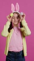 förtjusande liten flicka spelar titt en bua spel i studio, som visar henne handgjord färgad påsk ägg mot rosa bakgrund. glad lekfull unge med kanin öron lurar runt om. kamera b. video