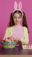 mignonne souriant bambin décorer une panier avec peint Pâques œufs, en présentant sa fabriqués à la main de fête arrangement dans studio. content charmant enfant création décorations et ornements. caméra b. video