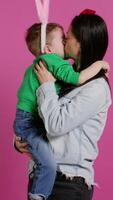 Cute mother and child showing love to each other in studio, mom hugging and kissing her small toddler son with bunny ears. Adorable little family laughing together and having fun. Camera B. video