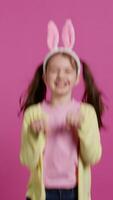 Joyful carefree schoolgirl jumping around in studio, imitating a rabbit and hopping against pink background. Cheerful active child wearing bunny ears and bouncing, adorable kid. Camera B. video