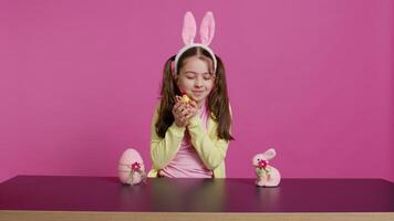 joyeux peu fille en jouant avec de fête Pâques décorations dans studio, création arrangements avec une poussin, lapin et œuf. souriant mignonne bambin avec lapin oreilles montrant coloré ornements. caméra b. video