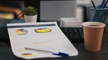 Financial report on desk next to paper cup of coffee in empty office, close up shot. Company files containing economic graphs and pie charts near hot beverage on table in workspace video