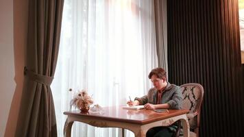 Man writing in notebook at a home office desk with curtains and natural light. video