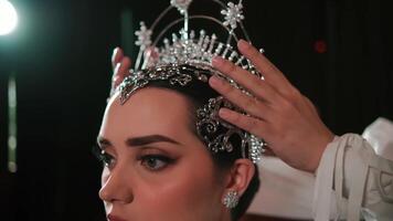 estilista ajustando uma tiara em uma jovem mulher cabeça, preparando para uma formal evento, com uma borrado fundo. video