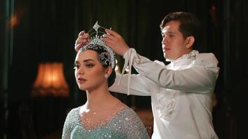 Elegant woman in a sparkling dress getting a tiara adjusted by a person in a white suit against a dark background. video