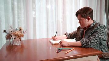 Man in casual attire writing in notebook at home with colored pencils on table, natural light from window. video