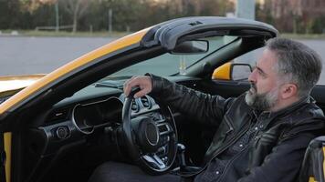 A confident man is sitting in a yellow sports car. Mature man with gray beard relaxing in a parked yellow convertible sports car, video