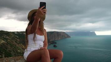 Woman travel sea. Happy tourist in hat enjoy taking picture outdoors for memories. Woman traveler posing on the beach at sea surrounded by volcanic mountains, sharing travel adventure journey video