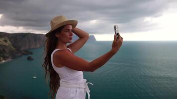 Woman travel sea. Happy tourist in hat enjoy taking picture outdoors for memories. Woman traveler posing on the beach at sea surrounded by volcanic mountains, sharing travel adventure journey video