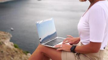 digital nômade, o negócio mulher trabalhando em computador portátil de a mar. bonita senhora digitando em computador de a mar às pôr do sol, faz uma o negócio transação conectados a partir de uma distância. freelance, controlo remoto trabalhos em período de férias video