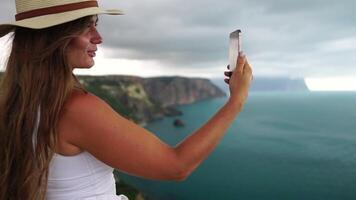 mujer viaje mar. contento turista en sombrero disfrutar tomando imagen al aire libre para recuerdos. mujer viajero posando en el playa a mar rodeado por volcánico montañas, compartiendo viaje aventuras viaje video