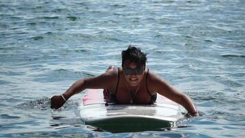 Happy body positive woman smiling and paddling on her surfboard in ocean or sea to beach after surfing on sunny day. Modern individual female hipster outdoor summer sport activity. Slow Motion video