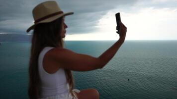 Woman travel sea. Happy tourist in hat enjoy taking picture outdoors for memories. Woman traveler posing on the beach at sea surrounded by volcanic mountains, sharing travel adventure journey video