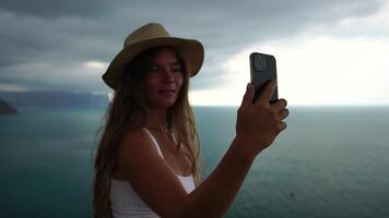 Woman travel sea. Happy tourist in hat enjoy taking picture outdoors for memories. Woman traveler posing on the beach at sea surrounded by volcanic mountains, sharing travel adventure journey video