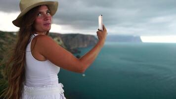 mujer viaje mar. contento turista en sombrero disfrutar tomando imagen al aire libre para recuerdos. mujer viajero posando en el playa a mar rodeado por volcánico montañas, compartiendo viaje aventuras viaje video