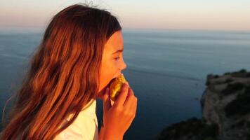 contento niña comiendo maíz. verano merienda en el mar. retrato de joven hermosa mujer comiendo A la parrilla maíz mientras sentado por el mar en puesta de sol tiempo. cerca arriba. lento movimiento. video