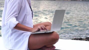 digital nômade, o negócio mulher trabalhando em computador portátil de a mar. bonita senhora digitando em computador de a mar às pôr do sol, faz uma o negócio transação conectados a partir de uma distância. freelance, controlo remoto trabalhos em período de férias video