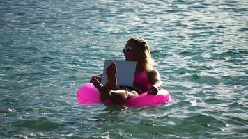 vrouw freelancer werken Aan laptop zwemmen in zee Aan roze opblaasbaar ring. mooi dame typen Aan computer terwijl drijvend in de zee Aan opblaasbaar donut Bij zonsondergang. freelance, afgelegen werk Aan vakantie video