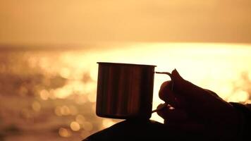 Travel stainless steel mug in the hands of a woman. Small waves on golden warm water surface with bokeh lights from sun. A concept of the world of beauty, nature and outdoor travel. Close up video