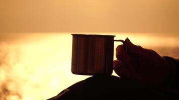 Travel stainless steel mug in the hands of a woman. Small waves on golden warm water surface with bokeh lights from sun. A concept of the world of beauty, nature and outdoor travel. Close up video