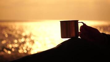 Travel stainless steel mug in the hands of a woman. Small waves on golden warm water surface with bokeh lights from sun. A concept of the world of beauty, nature and outdoor travel. Close up video