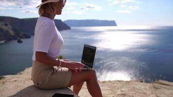 digital nômade, o negócio mulher trabalhando em computador portátil de a mar. bonita senhora digitando em computador de a mar às pôr do sol, faz uma o negócio transação conectados a partir de uma distância. freelance, controlo remoto trabalhos em período de férias video