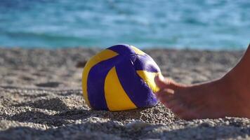 detailopname van een Mens schoppen een leeggelopen voetbal bal Aan de strand. een vent staand Aan de zand schopt een geel en blauw voetbal bal. langzaam beweging video