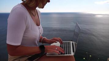 digital nômade, o negócio mulher trabalhando em computador portátil de a mar. bonita senhora digitando em computador de a mar às pôr do sol, faz uma o negócio transação conectados a partir de uma distância. freelance, controlo remoto trabalhos em período de férias video