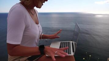 digitaal nomade, bedrijf vrouw werken Aan laptop door de zee. mooi dame typen Aan computer door de zee Bij zonsondergang, maakt een bedrijf transactie online van een afstand. freelance, afgelegen werk Aan vakantie video