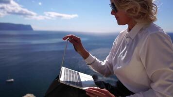 digitaal nomade, bedrijf vrouw werken Aan laptop door de zee. mooi dame typen Aan computer door de zee Bij zonsondergang, maakt een bedrijf transactie online van een afstand. freelance, afgelegen werk Aan vakantie video