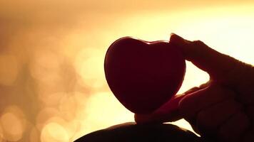 rojo corazones símbolo de amor en mano de mujer en el playa cerca el mar. amor concepto. pequeño olas en agua superficie en movimiento difuminar con dorado bokeh luces desde Dom. día festivo, vacaciones y recreación video