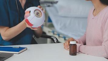 fechar acima do ásia fêmea médico vestindo face mascarar é mostrando globo ocular modelo e explicando sobre olhos doença para mais velho Senior homem paciente dentro hospital video