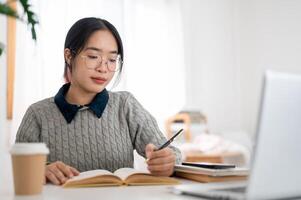 un joven asiático hembra estudiante es enfoque en su deberes y leyendo libro a un mesa en su habitación. foto