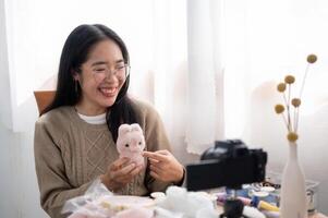 A smiling Asian female content creator is talking with the camera, showing her handmade plushie. photo