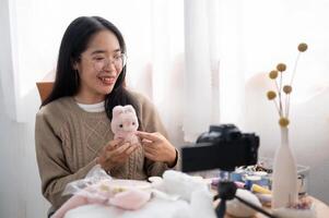 A smiling Asian female content creator is talking with the camera, showing her handmade plushie. photo