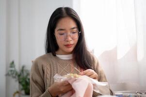 un joven asiático mujer es enfoque en enhebrar un modelo en un bordado marco, mano de coser en paño. foto