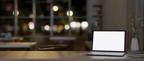 A table features a white-screen laptop computer mockup in a modern office at night. photo